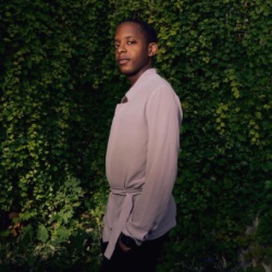 A person with short hair stands outdoors against a lush green wall of foliage. 他们穿着浅色的衣服, long-sleeve shirt and are looking slightly to 的 side with a calm expression. 的 lighting casts a soft shadow, creating a warm, natural ambiance.